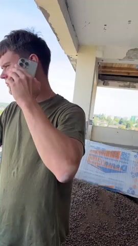 A man with short, dark hair is standing on what appears to be a balcony or unfinished building, wearing a green t-shirt and camouflage pants. He is holding a light-colored smartphone to his right ear and looking out at a view of houses and trees. His left arm is bent at the elbow, and his hand is resting on a concrete ledge in front of him. The sky is blue with some clouds. In the background, there is construction debris and materials.
