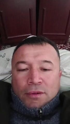 A close-up, head-and-shoulders shot shows a middle-aged man lying down, possibly in bed, as suggested by a pillow under his head. The background features a dark-colored wooden wardrobe. A small colorful toy can be seen on the right side. He appears to be speaking.
