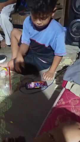 The video shows a group of children in what appears to be a dimly lit, indoor setting, possibly a home. In the foreground, a young boy with dark, curly hair is seated on the floor, engrossed in playing a mobile game on his smartphone. The phone is connected to a charger. He is wearing a blue and black t-shirt and black shorts. Next to him on the floor is a plastic cup with a red straw and a clear liquid. Further back, another boy in a green t-shirt and grey pants is seated on a wooden bench, looking in the direction of the boy playing the game. Other children are partially visible in the background. The environment is somewhat cluttered, with various items of clothing and furniture. A large speaker unit is visible on the right side of the frame. The overall atmosphere suggests a casual, informal gathering.