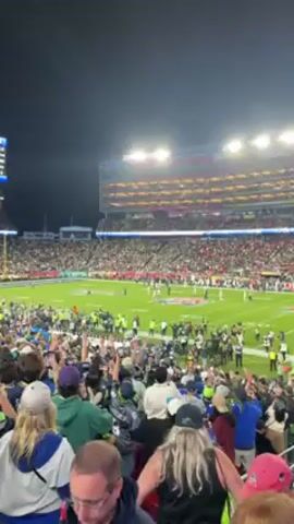 The video depicts a jubilant crowd in a packed stadium at night.  The spectators are mostly standing and raising their hands in celebration, their faces lit by the stadium lights.  They are wearing a variety of clothing, including sports jerseys and hats.  Below them, on the green field, figures can be seen, possibly players or staff, also celebrating.  The stadium is brightly lit and filled with spectators, creating a vibrant and energetic atmosphere.