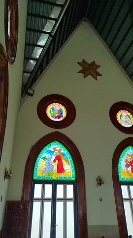 This media file captures the interior of a church in K'Rèn, Vietnam, viewed from a low angle looking upwards. The architecture features a distinct peaked roof with exposed metal beams and a tiled ceiling. The walls are painted a light color, adorned with decorative star-shaped elements and circular and arched stained-glass windows. These windows depict religious scenes with figures in colorful robes, suggesting biblical narratives.

The church is furnished with rows of dark wooden pews, arranged in a central aisle, indicative of a place of worship. The overall lighting suggests it could be daytime, with natural light filtering through the windows. The atmosphere appears serene and reverent, characteristic of a church setting.

One prominent feature is a large mural at the far end of the church, depicting a religious scene with multiple figures, possibly a crucifixion or ascension, rendered in a vibrant palette. Smaller alcoves with statues are also visible along the walls. The camera's perspective creates a sense of scale and grandeur, emphasizing the architectural details and decorative elements of the sacred space. No visible text is present in the media file.