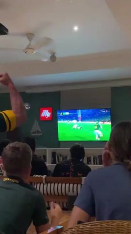 The video shows a group of people watching a rugby match on a large television screen. The room appears to be a living room or a lounge area, with a ceiling fan, a disco ball, and a large bookshelf in the background. The people are seated on couches and chairs, facing the television. One person in the foreground is wearing a green and yellow shirt, possibly a rugby jersey. Another person is wearing a black t-shirt and is gesturing with their hands, appearing to be excited or frustrated by the game. A man in the background is wearing a black t-shirt and a woman is sitting next to him. The atmosphere seems to be lively and engaged with the game. The television displays a rugby field with players in action. There is also an "Illy" coffee advertisement on the wall.