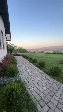 This is a vertical, wide shot of a cobblestone pathway leading into a green lawn with several neatly trimmed bushes and a small, red-leaved tree. In the background, there are fields, a fence, and mountains under a clear blue sky with a hint of sunset colors near the horizon. The scene appears to be set at dusk.