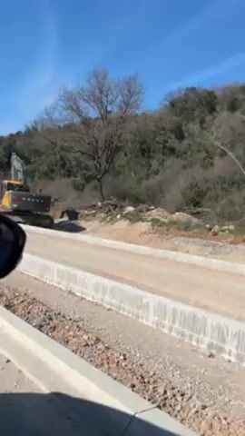 The video shows a yellow and black Volvo excavator parked on a dirt road. The road is bordered by new concrete curbing.  A small, leafless tree is in the background against a hillside covered in scrubby vegetation. The sky is blue and clear. The video is shot from inside a moving vehicle.
