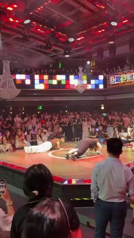 A group of people are performing a dance on a circular stage with a red and gold border. Two performers in the foreground are in a plank position. The audience is seated around the stage and in tiered seating above. The environment is a dimly lit venue with a large chandelier and colorful lights on the ceiling.