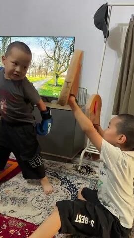 Two young children are playing indoors, possibly boxing. The child on the left is standing and wearing boxing gloves and black shorts, and appears to be punching towards a brown mitt held by the child on the right. The child on the right is sitting on the floor, also wearing shorts and holding the mitt. A television is visible behind the standing child, displaying a nature scene. The room has a carpeted floor and a clothing rack in the background.