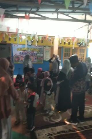 A group of people, likely students and teachers, are gathered in a room that appears to be a classroom or community space. They are all facing the same direction and are bowing, suggesting they are engaged in prayer. The room has a ceiling with exposed beams and is decorated with colorful flags and a banner that reads "SELAMAT DATANG TRIP CLASS". The floor is covered with a patterned rug. The lighting suggests it is daytime, possibly with sunlight streaming in from an unseen source. Some individuals are wearing headscarves, indicating a possible religious or cultural context for the gathering. The overall atmosphere seems to be one of communal activity and shared focus.