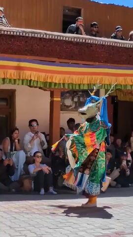A performer in a colorful, elaborate costume and mask with horns dances in a courtyard. Spectators are seated and standing around the perimeter, some holding cameras and phones. The performer's costume is predominantly green with various colorful patterns and embellishments. The background features traditional architecture with a balcony and colorful banners. The ground is paved with interlocking stones. The overall scene suggests a cultural performance or festival.