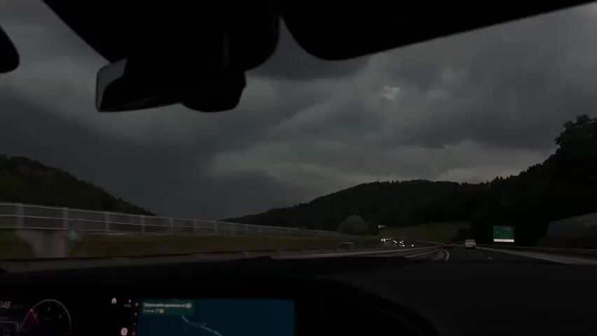 The video is taken from the interior of a car, looking forward through the windshield. It is daytime, but the sky is very dark and overcast, suggesting an impending storm. The car is on a highway, with other vehicles visible in the distance. The landscape outside consists of rolling hills covered in dark green trees. On the left side of the highway, there is a metal fence and some lighter green vegetation. A small, dark screen is visible in the lower left corner of the frame, likely a car's navigation or information display. The overall mood is dramatic due to the stormy weather.