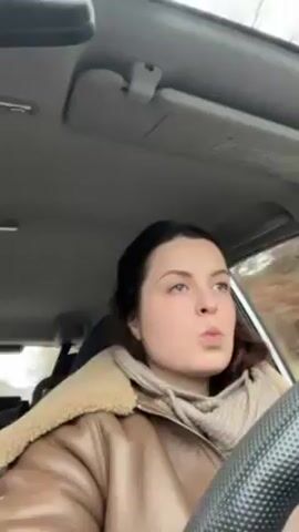 A young woman is driving a car. She's wearing a brown leather jacket with a sherpa collar and a beige turtleneck sweater. She has dark hair pulled back and blue eyes. The interior of the car is light gray. Music is playing in the background. The woman is looking at the road ahead and appears focused on driving. The environment outside the car appears to be a wooded area.