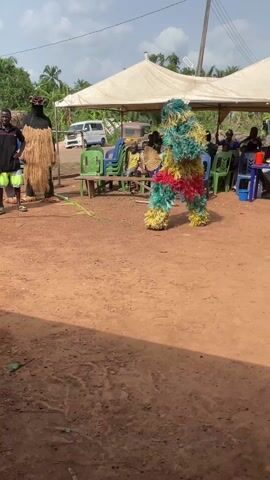 The scene features two costumed figures, one composed of light brown straw or fiber and the other more colorful, sporting green, yellow, and red sections. The latter dances in place, while three men stand nearby. They watch the costumed figures. In the background, under a white canopy, individuals sit on plastic chairs at a table. The ground is bare dirt, and vehicles are visible in the distance.