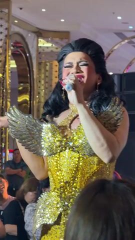 A drag performer, with dark hair styled in an updo, wears a bright gold, sequined dress with large, gold, feathered wings attached to the shoulders. The performer holds a microphone to their lips and sings, with their eyes closed and one hand placed over their chest. The performer has on heavy makeup, including dramatic eye makeup and bright red lipstick. They are on a stage in front of a crowd of people, some of whom are looking up at the performer. The background appears to be an indoor venue with decorative gold archways and lighting.