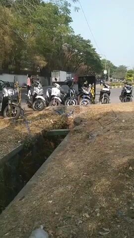 The video pans across a roadside scene. In the foreground, a deep, overgrown ditch runs horizontally. Beyond the ditch, several scooters and a three-wheeled vehicle are parked in a row along the side of a paved road. In the background, trees and foliage line the road, and the sky is visible above. There are some indistinct figures of people near the parked vehicles and in the distance, as well as some tents or market stalls. The overall environment appears to be outdoors in a moderately urban or suburban setting.