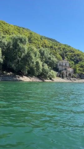 The video is a stationary, close-up shot of a body of water with a building and a forested hill in the background. The water in the foreground is a vibrant green with ripples. The building, which appears to be an old stone structure with arched windows and a domed roof, is situated on the shore of the water. The hill behind the building is covered in lush green trees and vegetation, with a clear blue sky above. The scene appears to be a serene and picturesque landscape.