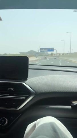 The video shows a car driving on a highway towards Zayed International Airport. The camera is positioned from the driver's seat, looking out through the windshield. A large blue sign overhead reads "Welcome to Zayed International Airport" with directions to Arrivals, Car Parks, Rental Return, and Departures. The highway appears to be relatively empty, with only a few cars visible in the distance. The sky is clear but slightly hazy, and the overall environment looks dry and desert-like. The car's dashboard is visible in the foreground, with a digital display and climate control buttons. The person holding the camera is wearing light-colored pants.