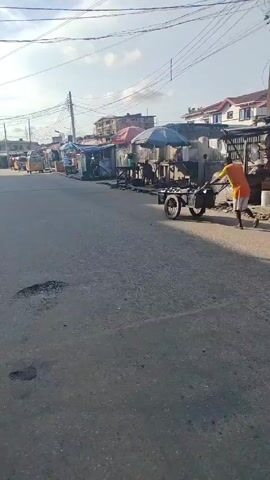The video shows a street scene in what appears to be a residential or commercial area.  The road is paved and has potholes. There are power lines overhead. Along the sides of the road, there are small shops and stalls. Several individuals can be seen in the video. A woman in a purple outfit with a shopping bag is walking along the road.  A man wearing an orange shirt is pulling a cart with what appears to be a person sitting on it.  A silver car drives along the road. The overall atmosphere suggests a typical day in a bustling neighborhood with some elements of commercial activity.