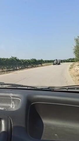 The video captures a view from inside a vehicle traveling on a paved road. The road curves to the right, bordered by vineyards on the left and olive trees and rocky terrain on the right. The sky is clear and blue. In the distance, a tractor with two people is visible on the road. Several black and white chevron road signs are placed along the curves of the road, warning of upcoming turns. The interior of the vehicle's dashboard is also visible in the foreground.