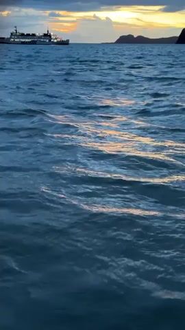 This is a video taken from the perspective of someone on a boat, looking out at a vast expanse of blue water. The water has gentle waves that reflect the light of the sky. In the distance, a large ferry can be seen moving across the water. The sky above is a mix of dark clouds and a warm, orange glow from what appears to be a sunset or sunrise. There is also a dark silhouette of land visible on the horizon behind the ferry. The overall mood of the video is serene and peaceful.