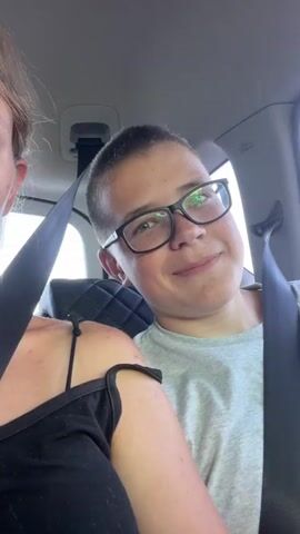 A short video clip captures two young people, a girl and a boy, sitting side-by-side in a car. The girl is positioned on the left, her face partially visible as she looks towards the camera with a neutral expression. She is wearing a black top with thin straps and a beaded necklace. The boy is on the right, closer to the camera, and he is smiling and looking directly at the viewer. He is wearing glasses and a light gray t-shirt. Both individuals are wearing seatbelts, indicating they are traveling in a vehicle. The interior of the car is visible, with a headrest behind the boy and a car seatbelt mechanism in the background. The lighting suggests it is daytime. The video appears to be a selfie taken by the girl or boy.