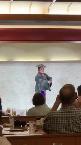 The video shows a traditional Japanese performance. A performer in a kimono and hat is dancing on a stage. There are people in the foreground watching the performance. The stage is lit with soft light, and the background is a textured wall. The performer is wearing a green and purple kimono with a white sash. The performer is moving gracefully, with fluid arm movements and a bent-knee stance. The audience members are seated at tables, and some are taking photos with their phones. The setting appears to be a restaurant or a cultural venue.