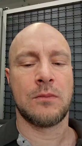 The video shows a close-up of a bald, middle-aged man with a beard. He is looking directly at the camera with a neutral expression. The background appears to be a metal grate. The man then smiles broadly, his eyes crinkling at the corners. He then puckers his lips and looks to the side, his eyes narrowed. He then looks back at the camera with wide eyes and a slightly open mouth, his expression one of surprise or curiosity.