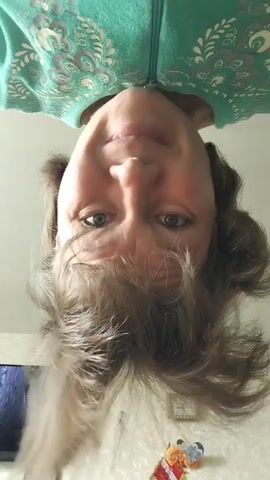 The video shows a close-up, upside-down shot of a woman's face as she leans towards the camera. She has blue eyes, light skin, and brown hair. She is wearing a teal hooded sweatshirt with a white floral pattern. Her mouth is open, and she appears to be speaking or making a sound. The background is out of focus but appears to be an indoor setting with a wall and possibly some objects on a surface.