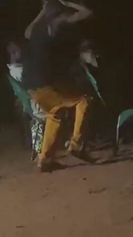 The video shows a person in yellow pants and a dark shirt dancing. They are bending at the waist, moving their arms, and turning. A green light is visible near them, and other people are in the background, though they are out of focus due to low lighting. The ground appears to be dirt or sand. The overall setting is dark and unclear.