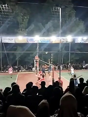 A night-time outdoor volleyball game is in progress. The court is lit by overhead lights. Spectators are seated in the foreground, silhouetted against the lights and the court. Players in red and blue uniforms are actively playing. The referee is positioned on a raised platform on one side of the net. The game appears to be competitive with players jumping and reaching for the ball. The atmosphere is energetic, with spectators watching intently.