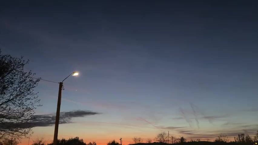 The video shows a nighttime scene with a streetlight in the foreground. The sky is a mix of dark blue and orange hues, suggesting it's dusk or dawn. There are thin clouds and contrails visible in the sky. Bare trees are silhouetted against the horizon. A child's voice can be heard, remarking on the brightness of the streetlight. The camera zooms in slightly on the streetlight.