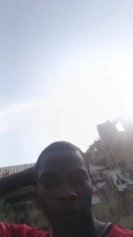 The video shows a young man with short, dark hair standing outdoors. He is wearing a red soccer jersey with the Manchester United logo and an Adidas logo on it. The camera appears to be hand-held, as it is close-up and captures the man's face and upper body. He repeatedly taps his head with his fist while looking at the camera. The background changes as the video progresses, showing different parts of what seems to be a residential area with some greenery.