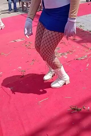 A medium shot focuses on the lower half of a person, likely female, dancing on a red carpet. The person is wearing white boots, fishnet stockings, a blue mini-skirt, and a white shirt with blue trim. The person also has on light-colored gloves and arm coverings. In the background, several people stand watching, and there are some structures and debris on the ground. The lighting appears to be natural daylight, casting shadows on the carpet.