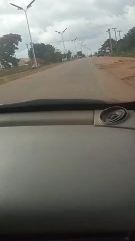The video shows a view from inside a car, looking forward through the windshield. The car is driving on a paved road lined with streetlights and trees. On either side of the road, there are buildings and other vehicles. The sky is cloudy. The camera is steady, indicating the car is moving at a constant speed. There are no people clearly visible in the foreground or engaging in any significant actions. The environment appears to be an urban or suburban area.
