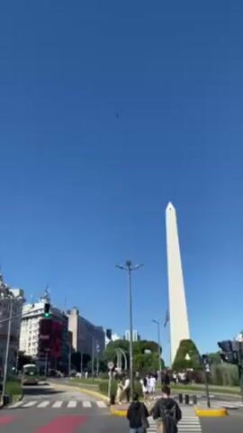 The video shows the Obelisco de Buenos Aires in Argentina on a bright, clear day. People are walking across the street in front of the monument. A few small objects, possibly birds, are flying in the bright blue sky above. The camera seems to be stationary and is positioned at street level.