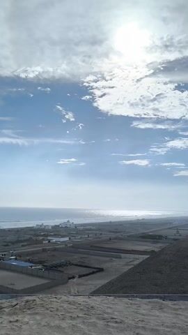 The camera pans across a sandy landscape with a view of the ocean. The sky is blue with white clouds, and the sun is shining brightly. The land in the foreground appears to be a construction site or an area with newly built structures, many rectangular enclosures or yards. Some buildings and sheds are visible on the land closer to the beach. The ocean stretches out to the horizon with gentle waves. The overall impression is of a coastal area under development on a sunny day.