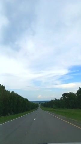 The video shows a view from a vehicle traveling on a paved road. The road stretches into the distance, bordered by a dense line of green trees on both sides. The sky is mostly filled with white clouds, with patches of blue visible. The scene appears to be a landscape view of a rural or undeveloped area. The camera is pointed forward, capturing the road ahead and the surrounding scenery. There are no people or other vehicles visible in the frame.