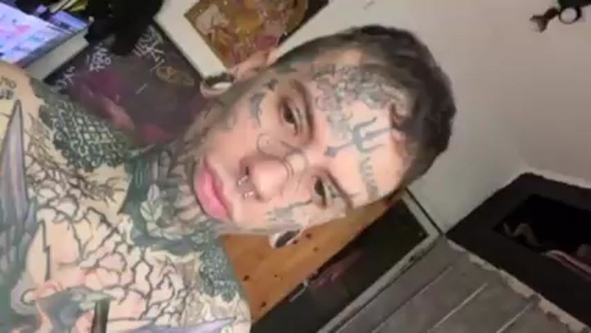 A person with extensive face and body tattoos is drinking from a glass jar with a metal straw. The jar appears to contain a leafy substance, possibly yerba mate. The person takes a sip, then briefly looks at the camera. The background appears to be a room with a door, a chair, and some items out of focus. The person is shirtless.