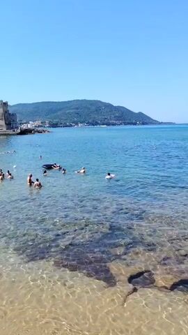 The video captures a sun-drenched beach scene with clear, shallow water lapping onto a sandy shore. The water is populated by numerous individuals, including adults and children, engaging in various aquatic activities. Some are swimming, while others are floating on inflatable rings and tubes. A small black boat is visible further out in the water. In the background, a historic stone building complex, resembling a castle or fortress, stands prominently along the coastline, with more buildings and a hillside covered in greenery extending beyond it. The sky is a clear, vibrant blue, indicating fair weather. The overall atmosphere appears relaxed and recreational, suggesting a popular tourist destination during summertime.