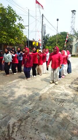 A group of women, most wearing red shirts and headscarves, are lined up outdoors. They appear to be participating in some kind of activity or event. The ground is paved, and there are trees and buildings in the background. A flag with red and white stripes is visible. The camera is held at an angle, making it difficult to determine the exact nature of the activity.