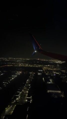The video shows an aerial view of a city at night, seen from the window of an airplane. The aircraft's wing is visible in the upper right corner of the frame. The city lights are spread across the horizon, showing streets and buildings. The lights are mostly white and yellow. The sky is dark, and a few stars are visible.