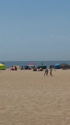 A wide shot shows a sandy beach with a clear blue sky. The ocean is visible in the background. Several people are scattered across the beach, some sitting in beach chairs or on towels, while others stand or walk. A few beach tents and umbrellas are set up. In the foreground, two young individuals, a boy and a girl, are walking towards the right side of the frame. The boy is wearing dark shorts and the girl is wearing a light-colored bathing suit. The overall scene depicts a sunny day at the beach with people enjoying leisure activities.