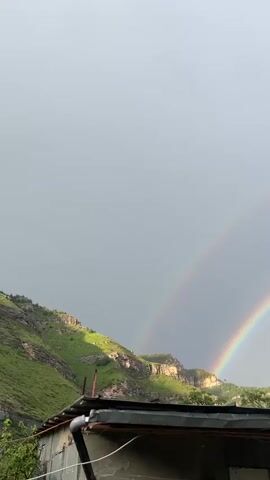 The video shows a cloudy sky with a double rainbow visible. In the foreground, the roof of a building is visible, with the side of a green, rocky mountain in the background. The image is oriented vertically.
