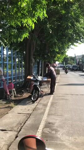 A man stands next to a motorcycle, which has a wooden table attached to its handlebars, with papers spread across it. He appears to be working with the papers, possibly organizing or folding them. The scene is outdoors, with trees lining the right side of the frame and a blue fence on the left. A sidewalk is visible in the foreground, and a road with a few vehicles can be seen in the background. The overall lighting suggests it is daytime.
