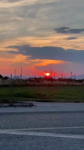 A video shows a sunset with orange and pink clouds over a field and a road. There are some power lines and a few distant structures visible in the background. The sun is a bright red orb just above the horizon.