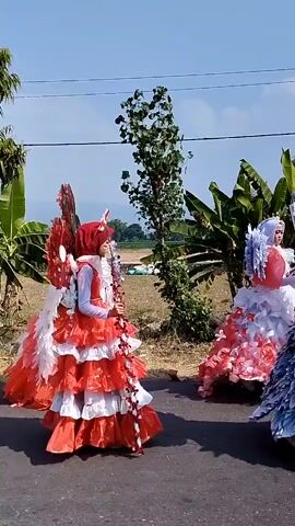 A group of people are walking down a paved road. The first person is wearing a red and white elaborate costume with large wings. The second person is wearing a similar costume. They are both walking on the right side of the road. Behind them, three people are walking on the left side of the road, all wearing hijabs and modest clothing. The environment appears to be rural with trees and fields in the background. The sky is clear and blue, suggesting it is daytime.