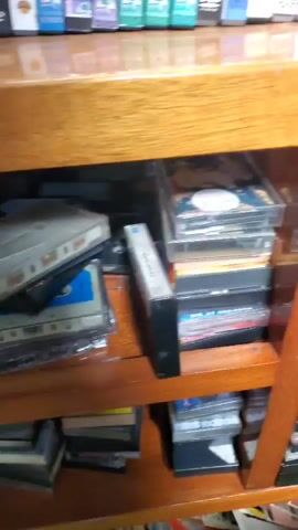 The camera pans across a bookshelf filled with various forms of media. On the top shelf are several DVDs with colorful spines. Below, the shelves are packed with cassette tapes and VHS tapes. The shelves themselves are made of a light-colored wood, possibly oak. In the final moments of the video, the camera focuses on a clear cassette tape.