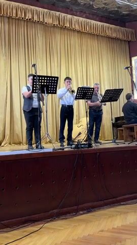 Three musicians are performing on a stage in an indoor setting, likely a hall or community center, in городское поселение Белогорск, Ukraine. The stage has a dark wooden front and golden curtains as a backdrop. Three men stand across the stage, each behind a music stand. The man on the left, wearing a vest over a white shirt, plays a large brass instrument, possibly a euphonium or baritone horn. The central musician, in a white shirt and black trousers, plays a trumpet. The man on the right, in a dark polo shirt, also plays a trumpet. All three are focused on their instruments and music sheets. Microphones on stands are positioned in front of them, and cables run across the stage floor, with a small black box labeled "VOTONE" visible. In the background to the right, a fourth musician is partially visible, seated and playing a piano. The overall atmosphere suggests a live musical performance.
