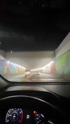 A car is driving through a tunnel with graffiti art on the walls. There are other cars in front of the car taking the video. The dashboard of the car is visible, showing that the car is in drive and the time is 8:51 PM. The tunnel is well-lit. The windshield of the car is slightly dirty.
