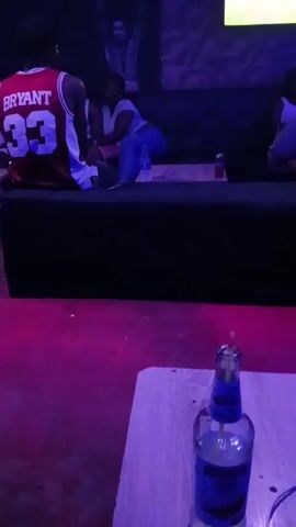 The camera is positioned low, focusing on a partially consumed beer bottle on a small table in the foreground. The room is dimly lit with blue and pink hues, creating a lounge-like atmosphere. In the background, several people are sitting on a black sofa. One person is wearing a red basketball jersey with the name "BRYANT" and the number 33 visible on the back. Two women are also sitting on the couch. The background features a textured wall with a picture or mural above the seating area and a television screen. The overall scene is a social gathering in a relaxed setting.