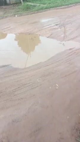 The video shows a muddy road with a large puddle in the center. A person in a blue shirt and patterned pants is walking away from the camera. There are buildings and overgrown vegetation on the sides of the road. The ground is wet and appears to have been recently rained on, with tire tracks visible in the mud. The overall environment suggests a rural or undeveloped area.