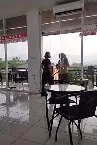 A video clip shows an interior view of a cafe or restaurant with a large glass window overlooking a misty, mountainous landscape. Inside, two women are standing near the window, appearing to converse. One woman is wearing a dark top and pants, while the other is wearing a light-colored top and brown pants with a headscarf. A round glass-topped table with two black chairs is in the foreground. The floor is tiled, and an air conditioning unit is mounted high on the wall. Banners with Indonesian text are visible on the left side of the window. The overall atmosphere suggests a calm, indoor setting with a scenic outdoor view.